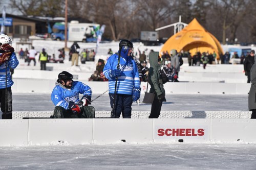 Two people on an ice rink, one sitting in a chair and the other standing, with a crowd and tents in the background.