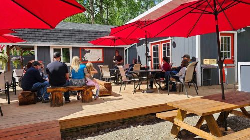 A group of people sitting at outdoor tables under red umbrellas, enjoying a sunny day at a cozy gathering space.