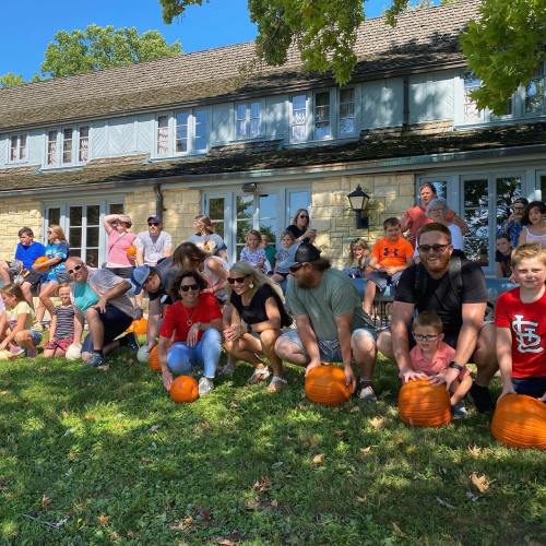 A large group of people sitting on a lawn with pumpkins in front of a stone house on a sunny day.