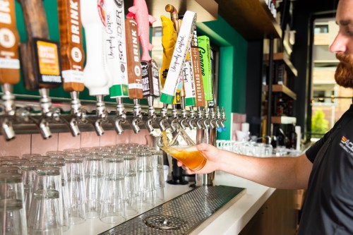 A bartender pours a drink from a row of colorful beer taps, with glasses lined up on the counter.