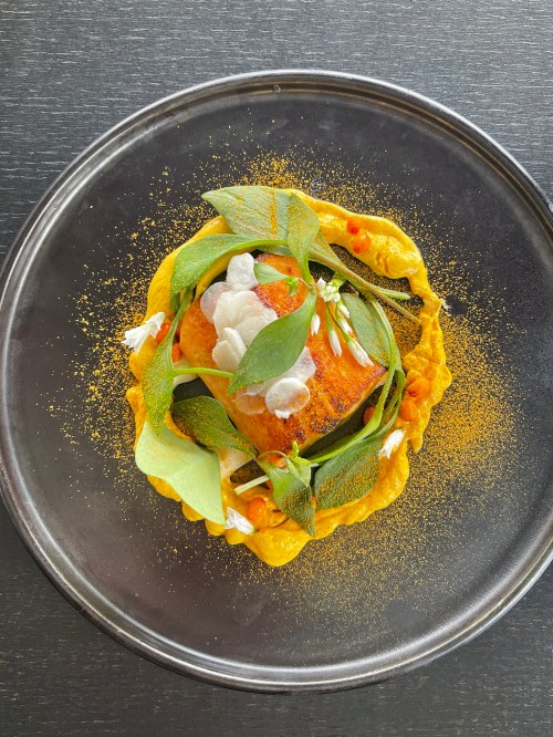 A beautifully plated dish featuring a piece of fish garnished with greens and edible flowers on a round metal plate.