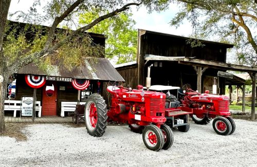 Two vintage red tractors parked outside a rustic garage adorned with patriotic decorations and surrounded by trees.