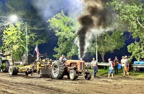 A vintage tractor pulls a heavy load, emitting black smoke, while spectators watch at a nighttime event.