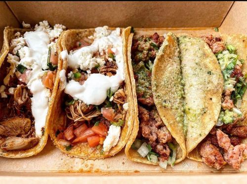Three tacos in a box: one with chicken, one with beef, and one with green sauce, topped with fresh ingredients.