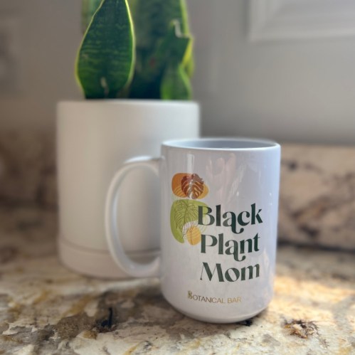 A white mug with "Black Plant Mom" text next to a green plant in a white pot on a countertop.