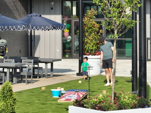 A man and a child play in a grassy area with outdoor seating and games, surrounded by trees and colorful balloons.