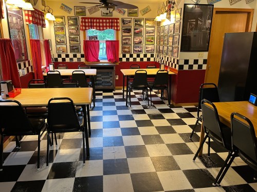 A cozy diner interior with checkered floors, black chairs, red curtains, and vintage car photos on the walls.