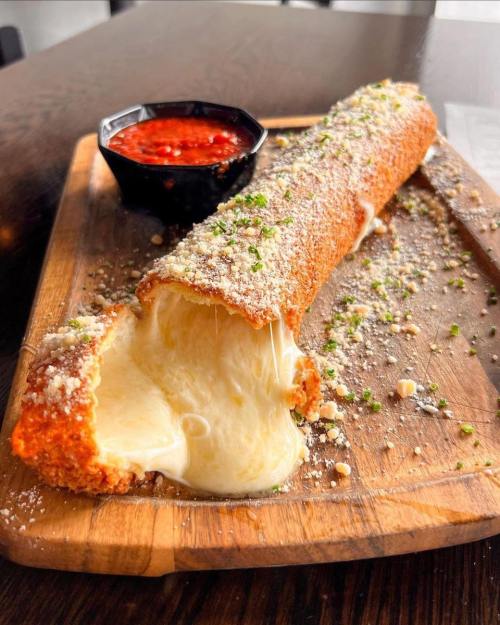 A crispy, cheese-filled roll on a wooden board, served with a side of marinara sauce and sprinkled with herbs.