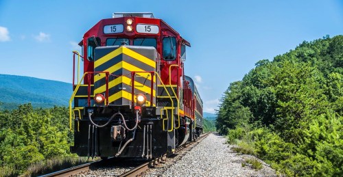 A red and yellow train on a track surrounded by green trees and mountains under a clear blue sky.