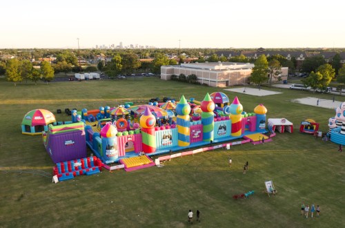 Aerial view of a colorful inflatable bounce park set up on a grassy field with city skyline in the background.