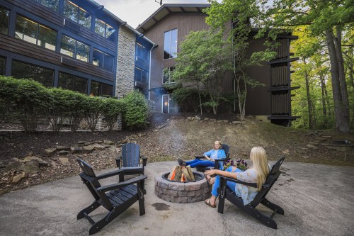 Two people relax by a fire pit surrounded by trees and a modern building, enjoying a cozy outdoor setting.