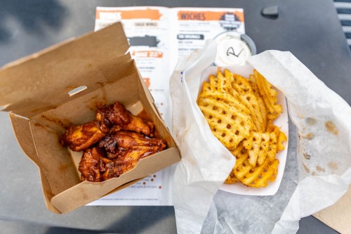 A takeout box with chicken wings next to a tray of waffle fries, placed on a table with a menu in the background.