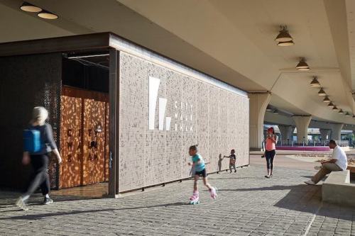 A modern building with a textured facade, people walking by, and a child on roller skates in a public space.