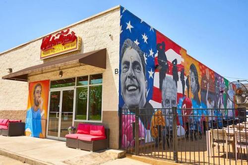Colorful mural on a restaurant wall featuring various portraits and an American flag backdrop. Outdoor seating visible.