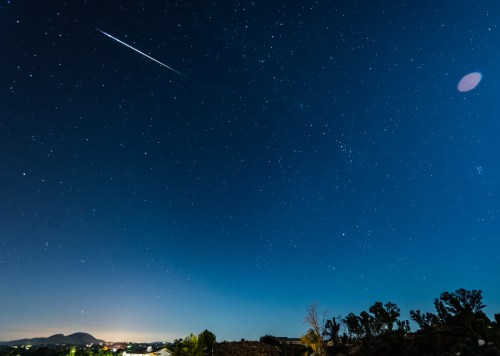 A clear night sky filled with stars, featuring a bright meteor streaking across the horizon above a distant landscape.