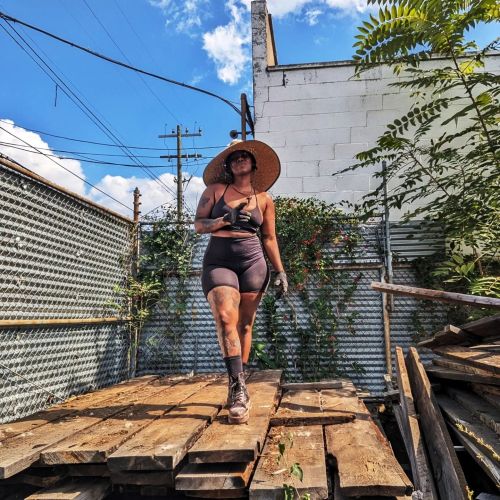 A person in a wide-brimmed hat walks on wooden planks in an outdoor setting, surrounded by greenery and buildings.
