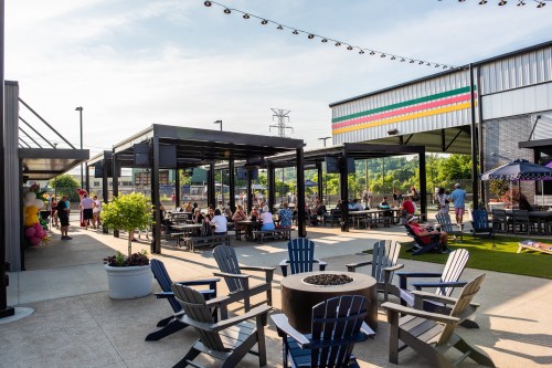 Outdoor gathering space with seating, fire pit, and people enjoying a sunny day at a lively venue.