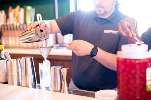 A bartender pours a cocktail through a strainer into a glass, with various bar tools and ingredients in the background.