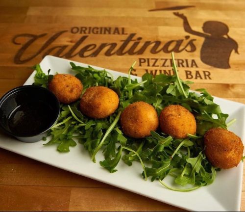 Four golden fried balls on a bed of arugula, served with a small cup of dipping sauce, against a wooden background.