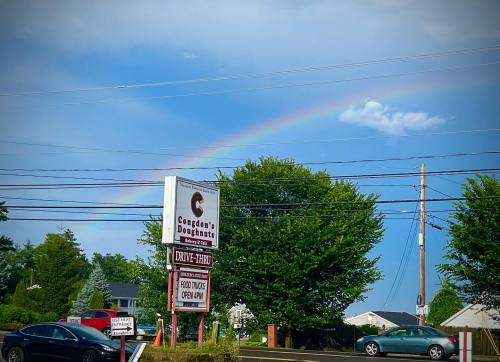 A sign for a doughnut shop under a rainbow, with trees and a blue sky in the background.