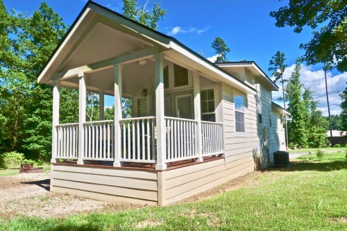 A small house with a porch, surrounded by trees and greenery under a clear blue sky.