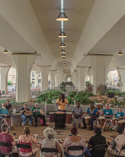 A diverse group of people playing drums in a spacious, open area under a large structure, with a woman leading the session.