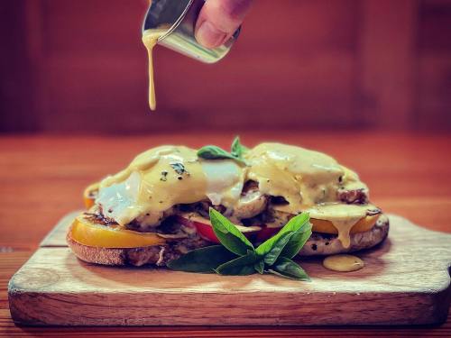 A hand drizzles sauce over a delicious open-faced sandwich topped with eggs, vegetables, and fresh basil on a wooden board.