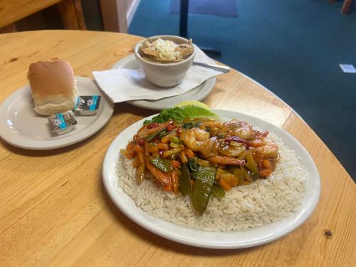 A plate of shrimp stir-fry with vegetables over rice, accompanied by a bowl of soup and a roll on a wooden table.