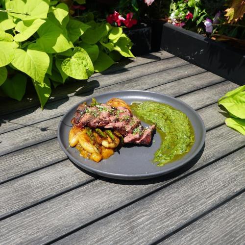 A plate of grilled steak with sautéed onions and a green sauce, set on a wooden table surrounded by greenery.
