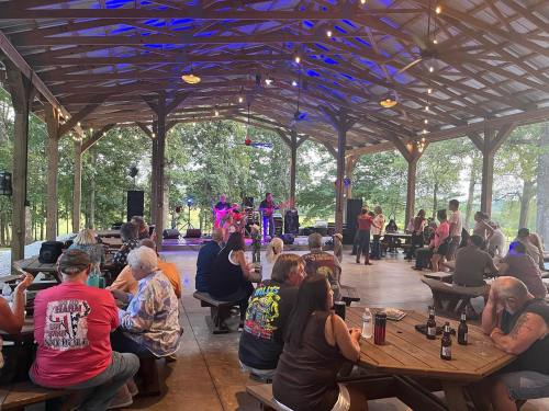 A lively outdoor gathering with a band performing under a wooden pavilion, surrounded by seated attendees enjoying the music.