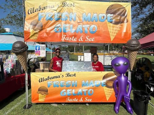 Two vendors stand behind a colorful gelato stand, featuring a large sign that reads "Fresh Made Gelato" and an alien mascot.