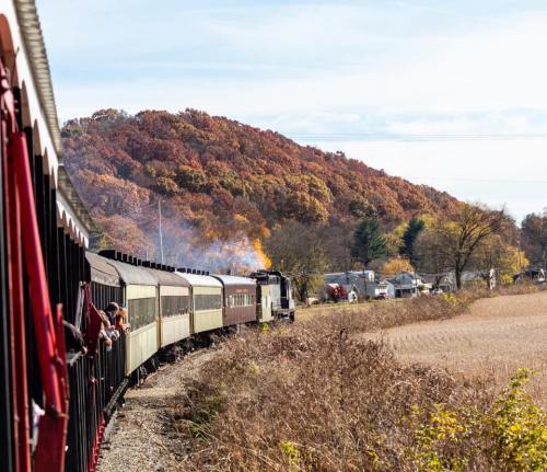 A vintage train winds through a scenic landscape with autumn foliage and a clear sky.