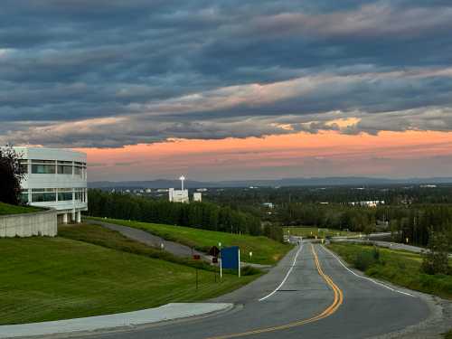 A winding road leads through green fields, with a modern building on the left and a colorful sunset in the background.