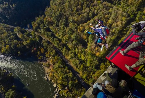 Aerial view of a person base jumping off a cliff into a river, surrounded by trees and spectators on the edge.