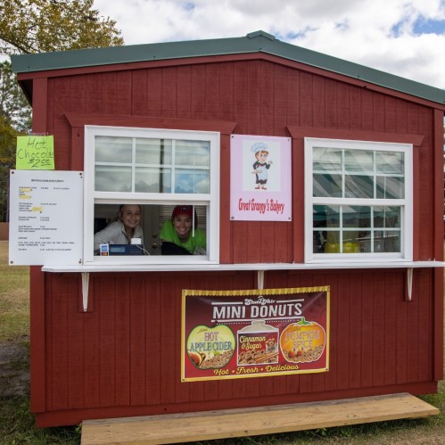 A small red food stand with two smiling employees inside, offering mini donuts and hot chocolate.