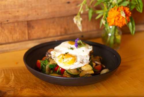 A bowl of sautéed vegetables topped with two fried eggs, with a small vase of flowers in the background.