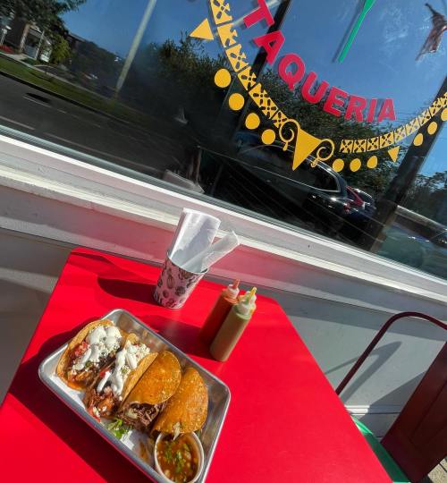 A vibrant taqueria scene featuring a plate of tacos, salsa, and condiments on a red table, with a window view.