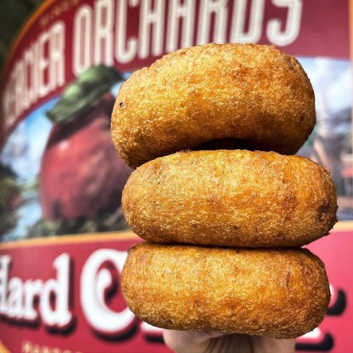 Three golden-brown donuts stacked on top of each other, with a blurred background featuring a fruit orchard sign.