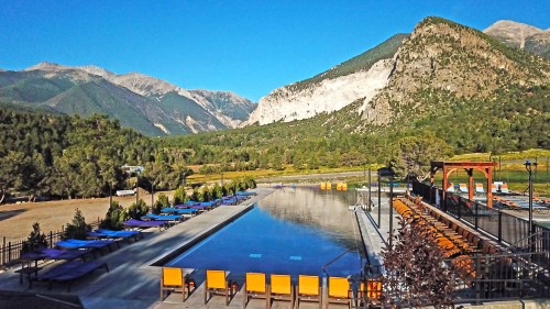 A serene poolside view with orange loungers, surrounded by mountains and lush greenery under a clear blue sky.