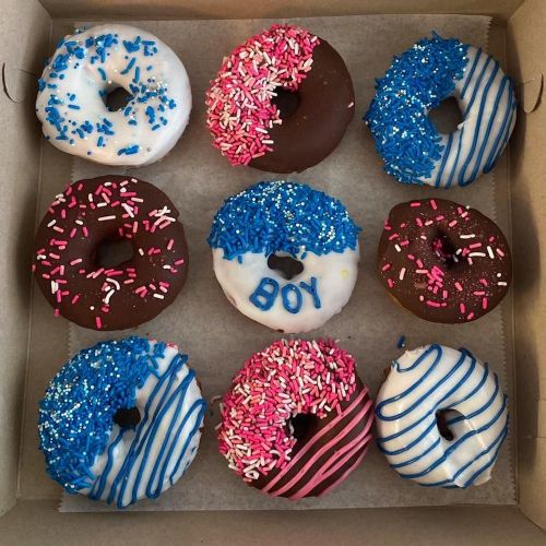 A box of nine donuts with various toppings, including blue and pink sprinkles, some decorated with the word "BOY."