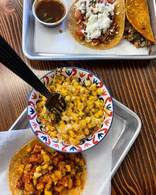 A colorful spread of tacos and a bowl of cheesy corn on a wooden table, with a side of salsa.