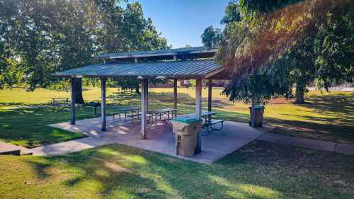 A shaded picnic area with tables and trash bins in a grassy park surrounded by trees.