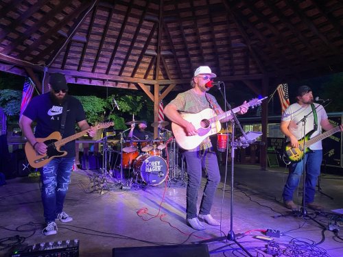 A band performs on stage under a pavilion, with a guitarist, bassist, and drummer, creating a lively atmosphere.
