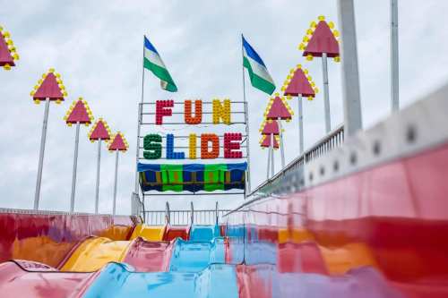 Colorful "FUN SLIDE" sign atop a vibrant slide with flags, set against a cloudy sky.