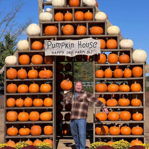 A man stands in front of a colorful display of pumpkins at a farm, with a sign reading "Pumpkin House."