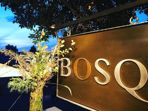 A wooden sign reading "BOSQ" with a vase of greenery in the foreground against a twilight sky.