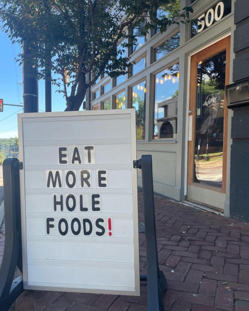 A sidewalk sign outside a store reads "EAT MORE HOLE FOODS!" with a sunny street view in the background.