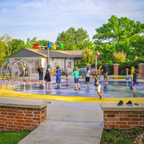 Children playing in a splash pad area with colorful water features and greenery in the background on a sunny day.