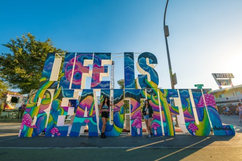 Two people pose in front of colorful "LIFE IS BEAUTIFUL" letters at an outdoor festival. Bright blue sky in the background.