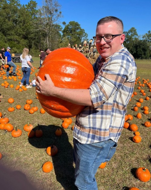 A person in a plaid shirt smiles while holding a large orange pumpkin in a pumpkin patch filled with smaller pumpkins.